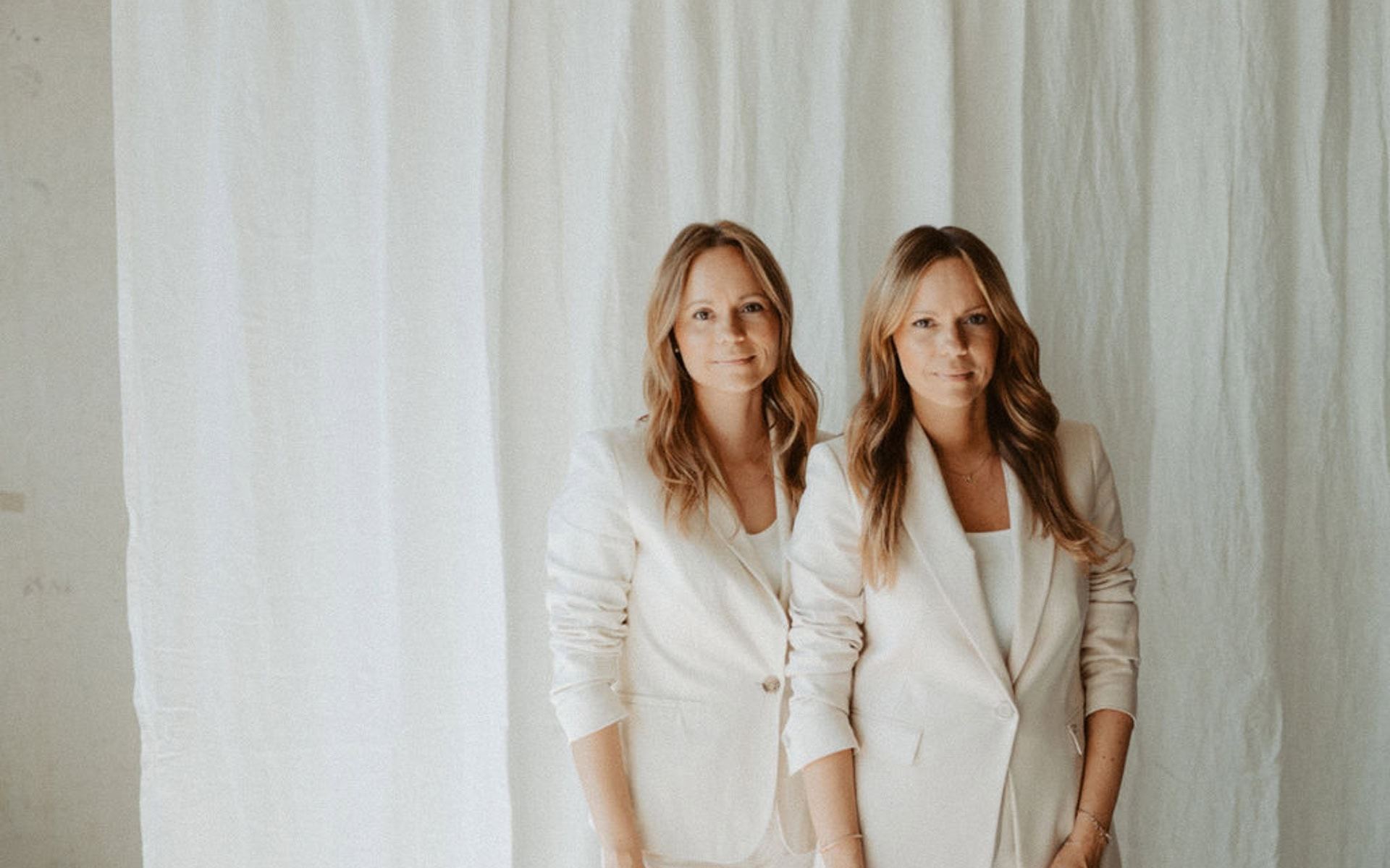 Pia und Nina, die Zwillingsschwestern hinter der Online Boutique Agentur, in hellen Anzügen vor einem Leinenvorhang