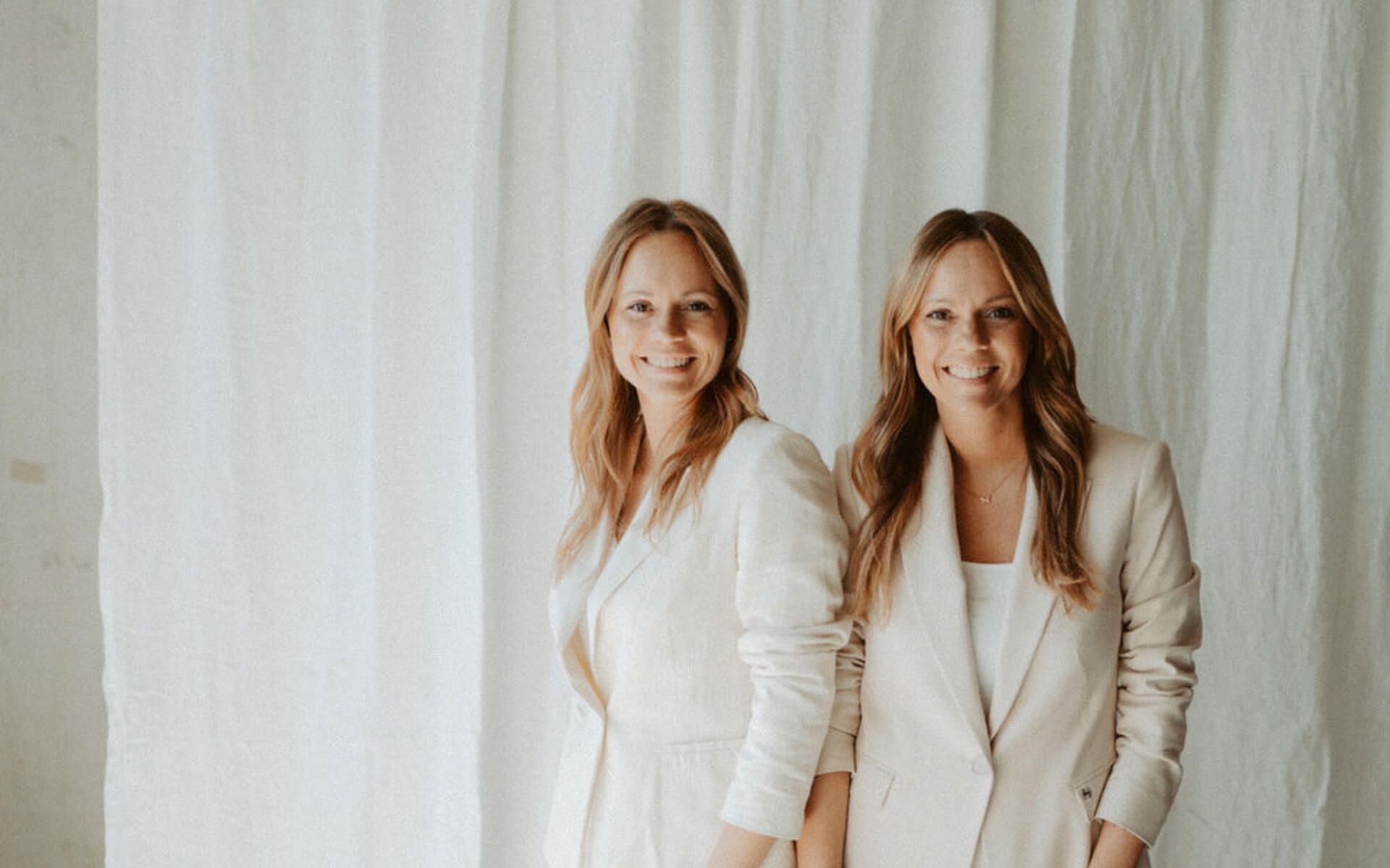 Nina und Pia, die Gründerinnen der Online Boutique Agentur, lachend in hellen Blazern vor einem Leinenvorhang